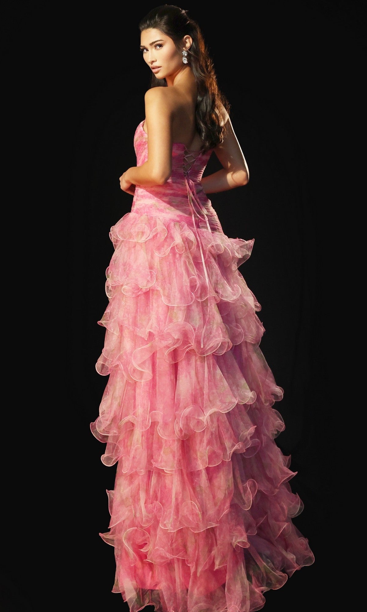 A woman stands against a black background wearing the Jovani 46408 long prom dress in watercolor pink, its strapless, floor-length ruffle organza gown cascading as she looks over her shoulder.
